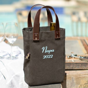 May include: A brown canvas wine tote bag with leather handles. The bag has a pocket on the side and is embroidered with the text "Napa 2022".