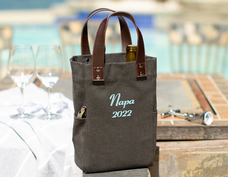 May include: A brown canvas wine tote bag with leather handles. The bag has a pocket on the side and is embroidered with the text "Napa 2022".