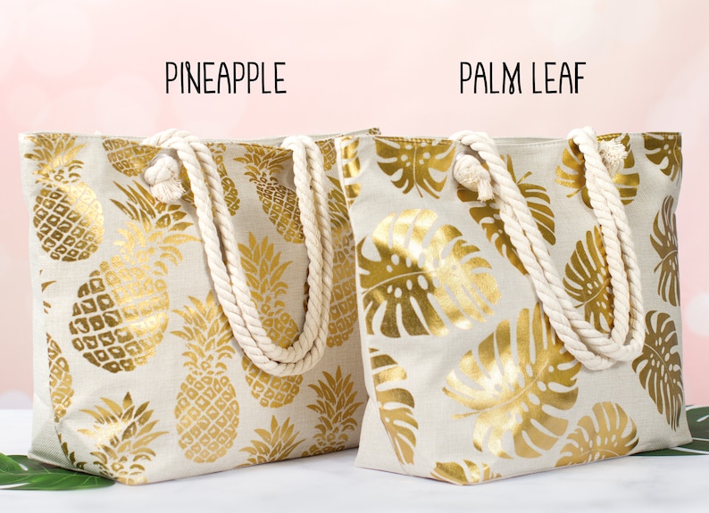 May include: Two canvas tote bags with rope handles. The bag on the left has a gold pineapple pattern on a beige background. The bag on the right has a gold palm leaf pattern on a beige background. The text "Pineapple" is above the left bag and the text "Palm Leaf" is above the right bag.