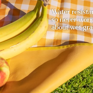 Puede incluir: Una manta de picnic a cuadros amarillos y blancos con un borde amarillo. La manta est&aacute; parcialmente extendida sobre hierba verde. Dos pl&aacute;tanos y dos manzanas rojas est&aacute;n sobre la manta. El texto en la manta dice: &iexcl;Resistente al agua, as&iacute; que nunca te preocupes por la hierba mojada!