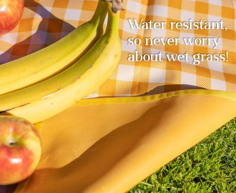 Puede incluir: Una manta de picnic a cuadros amarillos y blancos con un borde amarillo. La manta est&aacute; parcialmente extendida sobre hierba verde. Dos pl&aacute;tanos y dos manzanas rojas est&aacute;n sobre la manta. El texto en la manta dice: &iexcl;Resistente al agua, as&iacute; que nunca te preocupes por la hierba mojada!