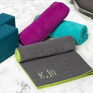 May include: Three yoga towels rolled up on a white marble surface. The towels are gray, teal, and purple. The gray towel has a green border and the initials "K.J.Q" embroidered in green. Two teal yoga blocks are also visible.