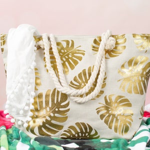 May include: A white canvas tote bag with gold leaf tropical leaf print and rope handles. The bag is sitting on a green and red patterned beach towel.