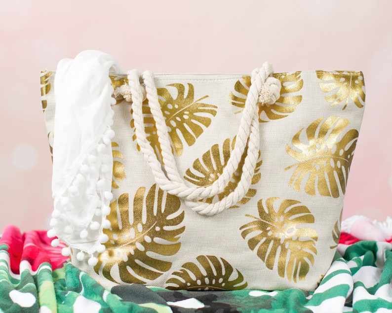 May include: A white canvas tote bag with gold leaf tropical leaf print and rope handles. The bag is sitting on a green and red patterned beach towel.