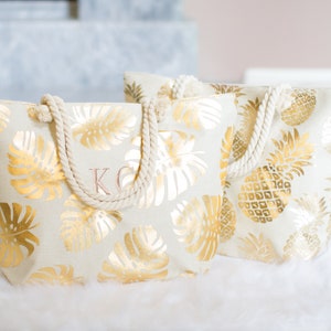 May include: Two white canvas tote bags with gold foil pineapple and monstera leaf patterns. The bag on the left has the initials "KC" embroidered on the front.