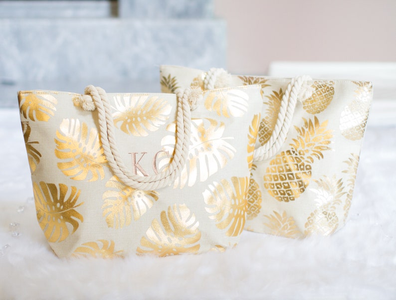 May include: Two white canvas tote bags with gold foil pineapple and monstera leaf patterns. The bag on the left has the initials "KC" embroidered on the front.