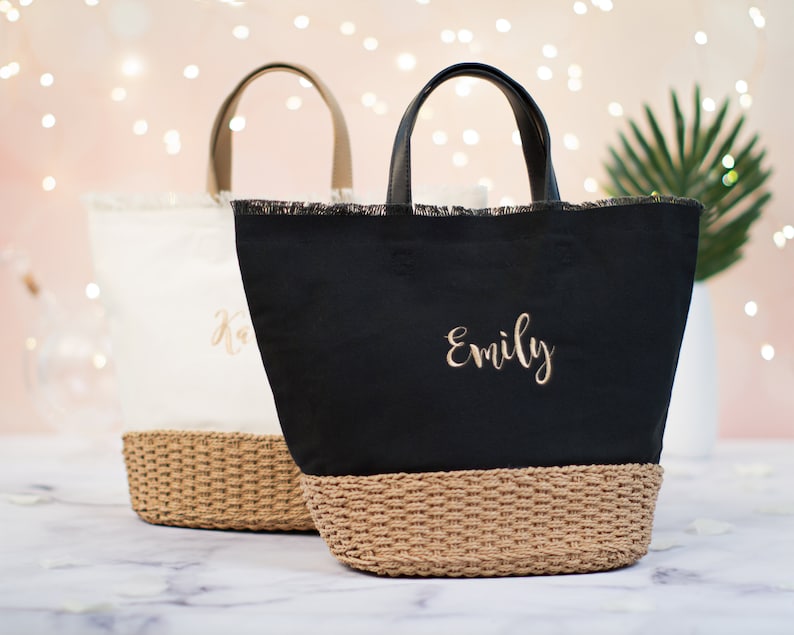May include: Two personalized tote bags with woven straw bottoms. The bag on the left is white with the name "Kara" embroidered in gold. The bag on the right is black with the name "Emily" embroidered in gold.