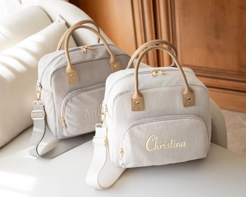 May include: Two light gray corduroy baby bags with tan handles and gold hardware. One bag has the name "Amelia" embroidered, the other "Christina". Both have a shoulder strap and a front pocket.