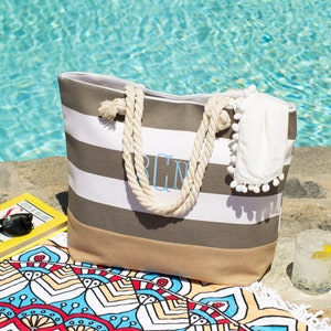 May include: A striped beach tote bag with rope handles, personalized with light blue initials. The bag is gray and white striped with a tan bottom. A white towel with pom-poms, sunglasses, and a book are nearby.