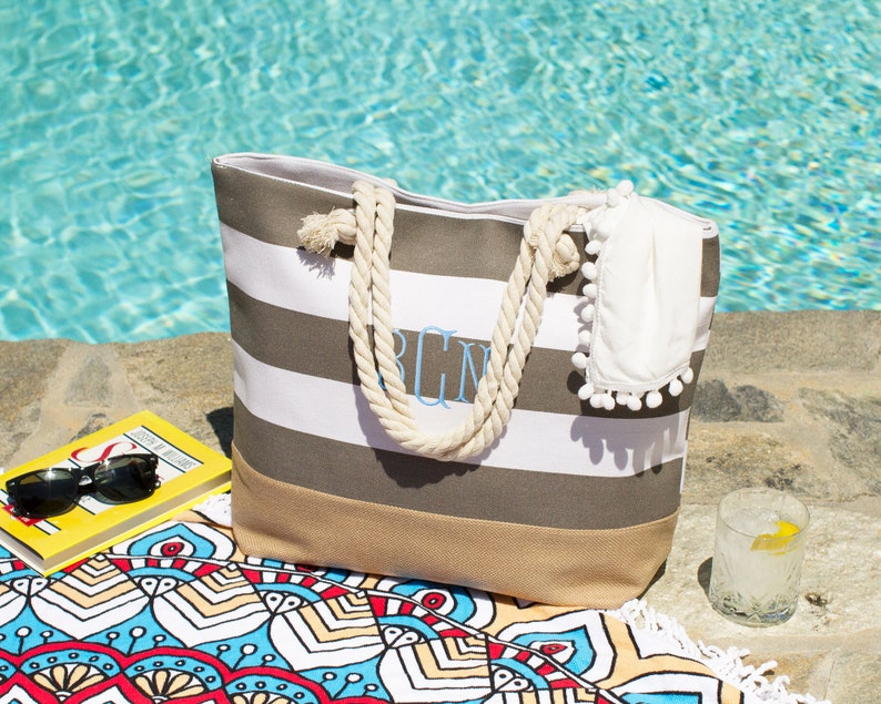 May include: A striped beach tote bag with rope handles, personalized with light blue initials. The bag is gray and white striped with a tan bottom. A white towel with pom-poms, sunglasses, and a book are nearby.
