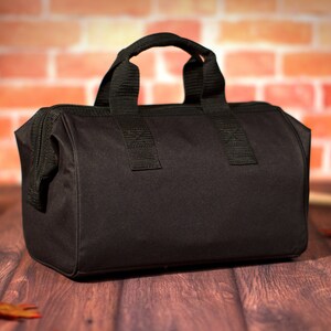 K&ouml;nnte beinhalten: Eine schwarze Werkzeugtasche aus Canvas mit Rei&szlig;verschluss und zwei Tragegriffen. Die Tasche liegt auf einer Holzoberfl&auml;che.