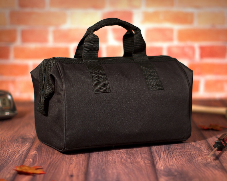 K&ouml;nnte beinhalten: Eine schwarze Werkzeugtasche aus Canvas mit Rei&szlig;verschluss und zwei Tragegriffen. Die Tasche liegt auf einer Holzoberfl&auml;che.