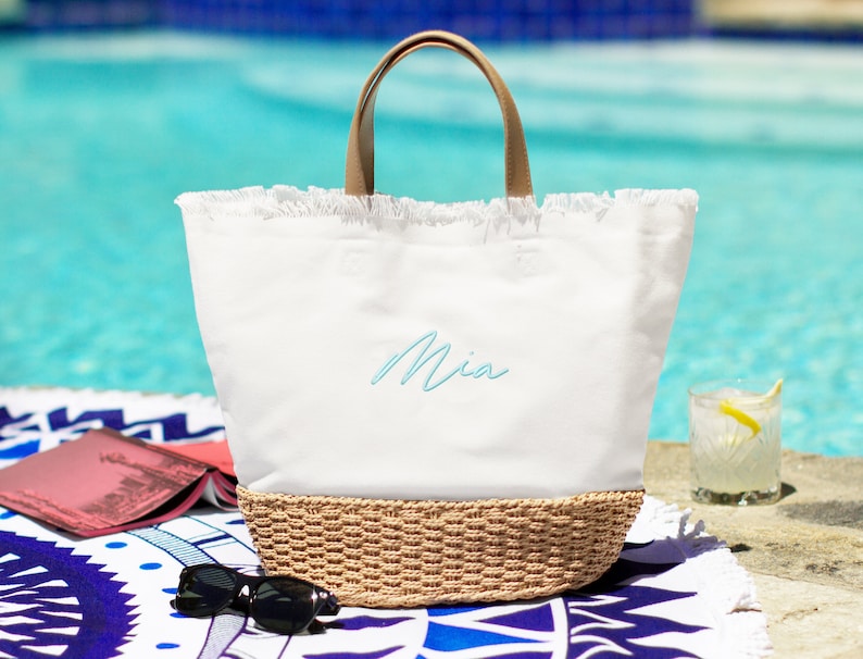 May include: White canvas tote bag with a brown leather handle and a woven straw bottom. The bag has a frayed edge and the name "Mia" is embroidered on the front in light blue thread.