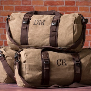 K&ouml;nnte beinhalten: Zwei braune Canvas-Reisetaschen mit braunen Lederelementen und schwarzen Riemen. Die obere Tasche hat die Initialen "DM" auf der Vorderseite gestickt. Die untere Tasche hat die Initialen "CR" auf der Vorderseite gestickt.