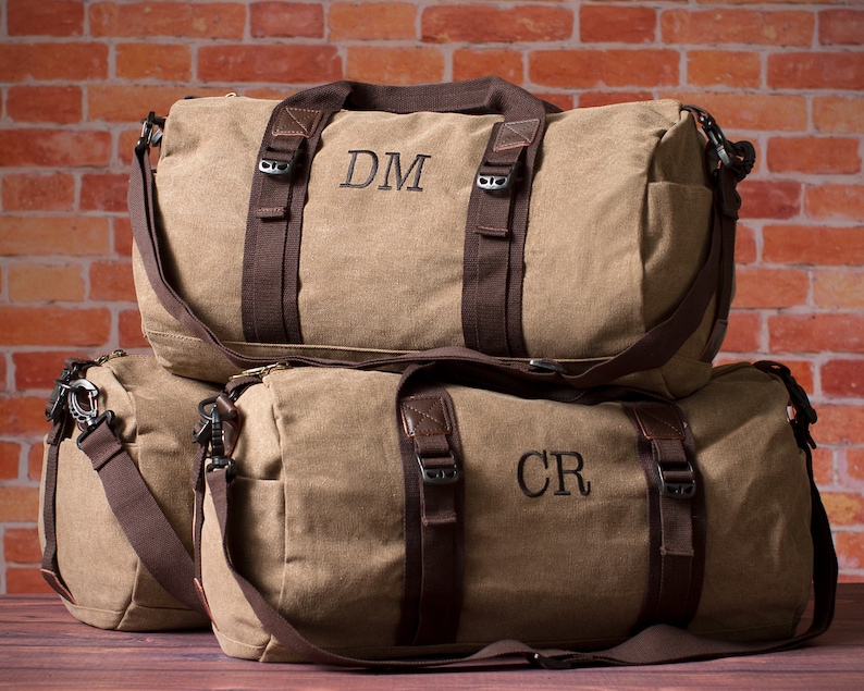 K&ouml;nnte beinhalten: Zwei braune Canvas-Reisetaschen mit braunen Lederelementen und schwarzen Riemen. Die obere Tasche hat die Initialen "DM" auf der Vorderseite gestickt. Die untere Tasche hat die Initialen "CR" auf der Vorderseite gestickt.