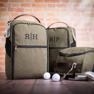 May include: Three green canvas golf shoe bags with black trim and leather accents. The bags have zipper closures and are monogrammed with the initials "R|H", "S|P", and a third bag with no visible initials. Two golf balls and golf clubs are in the foreground.