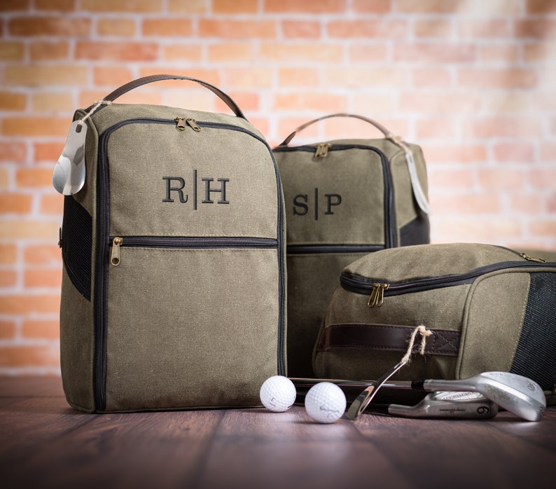 May include: Three green canvas golf shoe bags with black trim and leather accents. The bags have zipper closures and are monogrammed with the initials "R|H", "S|P", and a third bag with no visible initials. Two golf balls and golf clubs are in the foreground.