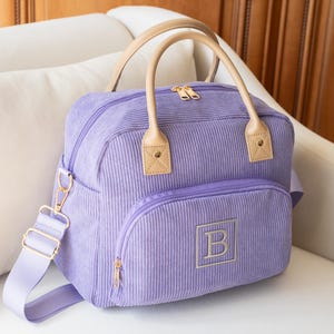 May include: A lavender corduroy bag with a top handle and a detachable shoulder strap. The bag features a front pocket with a gold zipper and an embroidered "B" in a square. The handles and accents are tan.