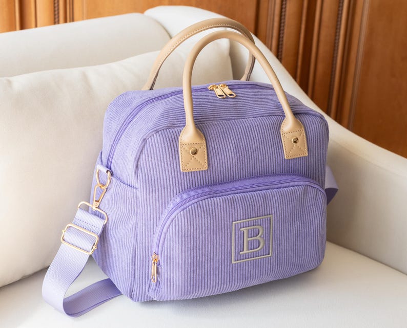 May include: A lavender corduroy bag with a top handle and a detachable shoulder strap. The bag features a front pocket with a gold zipper and an embroidered "B" in a square. The handles and accents are tan.
