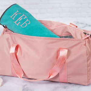 May include: A pink duffel bag with pink straps and a zipper closure. A teal towel with the monogram "K.E.B." is inside the bag.