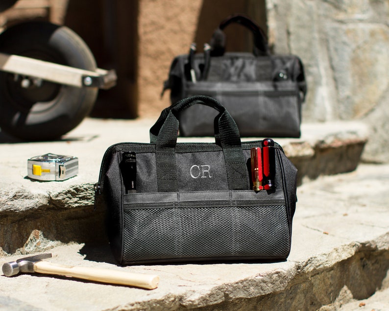 May include: Two black canvas tool bags with a zipper closure and multiple pockets. The bag on the left has the initials 'CR' embroidered on the front. The bag on the right has a red multi-tool sticking out of the side pocket.