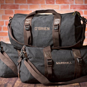 K&ouml;nnte beinhalten: Zwei schwarze Canvas-Reisetaschen mit braunen Lederakzenten und Riemen. Die gr&ouml;&szlig;ere Tasche hat den Namen "O'BRIEN" auf der Vorderseite gestickt. Die kleinere Tasche hat den Namen "MARSHALL" auf der Vorderseite gestickt.