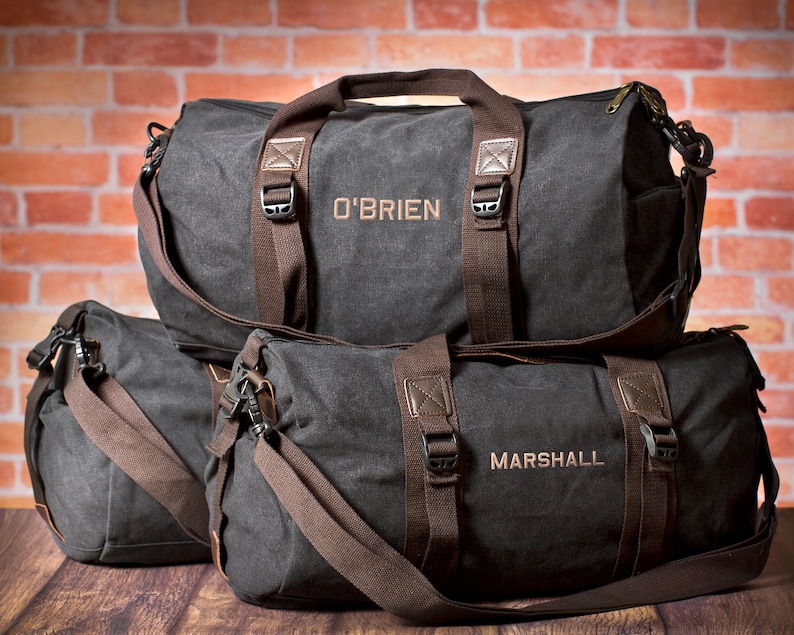 K&ouml;nnte beinhalten: Zwei schwarze Canvas-Reisetaschen mit braunen Lederakzenten und Riemen. Die gr&ouml;&szlig;ere Tasche hat den Namen "O'BRIEN" auf der Vorderseite gestickt. Die kleinere Tasche hat den Namen "MARSHALL" auf der Vorderseite gestickt.