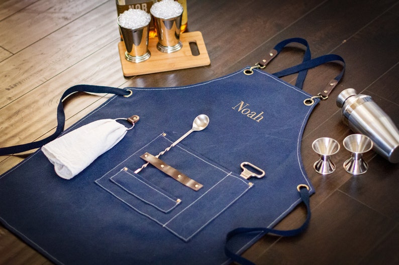 May include: A blue denim bar apron with a brown leather strap and a large pocket. The apron has a name embroidered on it, "Noah". The apron is laying on a wooden surface with a cocktail shaker, two jiggers, a bar spoon, and a bottle opener.