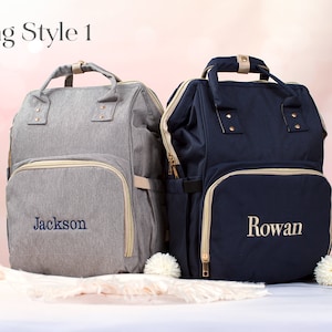 May include: Two gray and navy blue diaper bags with gold zippers and trim. The gray bag has the name "Jackson" embroidered on it, and the navy blue bag has the name "Rowan" embroidered on it.