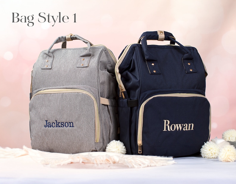 May include: Two gray and navy blue diaper bags with gold zippers and trim. The gray bag has the name "Jackson" embroidered on it, and the navy blue bag has the name "Rowan" embroidered on it.