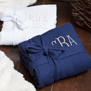 May include: A white waffle-weave robe with the embroidered monogram MKE and a navy blue waffle-weave robe with the embroidered monogram CRA. Both robes are folded neatly and feature a matching fabric tie belt wrapped around the center.