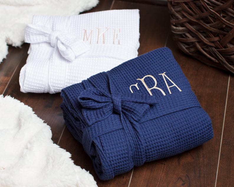 May include: A white waffle-weave robe with the embroidered monogram MKE and a navy blue waffle-weave robe with the embroidered monogram CRA. Both robes are folded neatly and feature a matching fabric tie belt wrapped around the center.