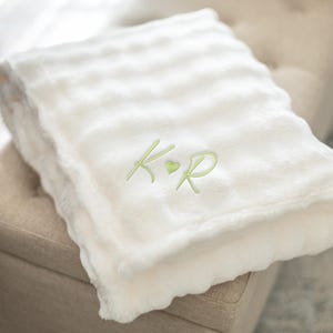 May include: A folded, cream-colored plush blanket with a scalloped edge. The blanket has a ribbed texture and is embroidered with the initials "K R" in a light green color, with a small green heart between the letters. The blanket is folded on a beige ottoman.