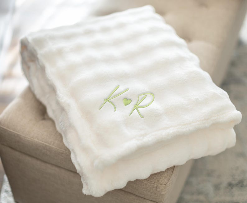 May include: A folded, cream-colored plush blanket with a scalloped edge. The blanket has a ribbed texture and is embroidered with the initials "K R" in a light green color, with a small green heart between the letters. The blanket is folded on a beige ottoman.