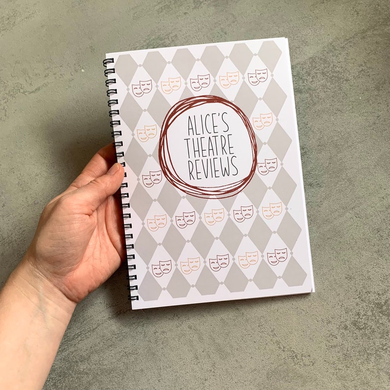 Puede incluir: Un cuaderno con espiral con una cubierta con un patr&oacute;n de diamantes grises y blancos y m&aacute;scaras de teatro. El centro de la cubierta tiene un c&iacute;rculo marr&oacute;n con el texto "ALICE'S THEATRE REVIEWS".