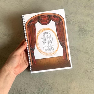 Puede incluir: Un cuaderno con espiral con una portada tem&aacute;tica de teatro. La portada presenta un escenario con cortinas rojas y el texto "AMY'S DAY OUT TO THE THEATRE" en un dise&ntilde;o circular. El cuaderno est&aacute; sostenido por una mano.