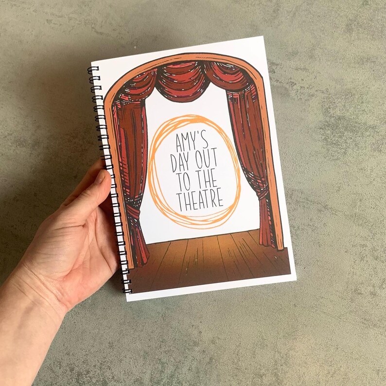 Puede incluir: Un cuaderno con espiral con una portada tem&aacute;tica de teatro. La portada presenta un escenario con cortinas rojas y el texto "AMY'S DAY OUT TO THE THEATRE" en un dise&ntilde;o circular. El cuaderno est&aacute; sostenido por una mano.