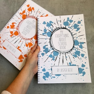 May include: Two spiral-bound notebooks with white covers featuring ink splatter designs. One cover has orange splatters and the text "MY EVERYTHING ABOUT SCHOOL BOOK BY MILO BECK," while the other has blue splatters and "MY EVERYTHING ABOUT SCHOOL BOOK BY JASPER BECK."