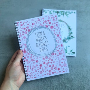 May include: A spiral-bound notebook with a white cover featuring a pattern of pink watercolour-style dots. Centred on the cover is a grey hand-drawn circle containing the black text: LEON & HONEY'S ALPHABET DATES. A second notebook is visible behind it.