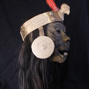 May include: A dark brown mask with a gold headband and a red feather. The mask has a bird beak and a round, white, textured ear ornament.