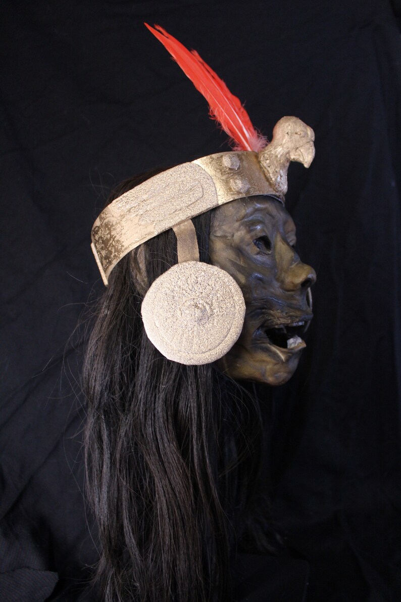 May include: A dark brown mask with a gold headband and a red feather. The mask has a bird beak and a round, white, textured ear ornament.