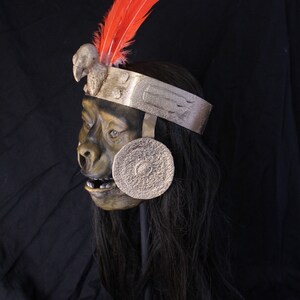 May include: A detailed, hand-crafted mask of a monkey wearing a silver headdress with red feathers. The mask has a realistic appearance with a textured, green face and long black hair.