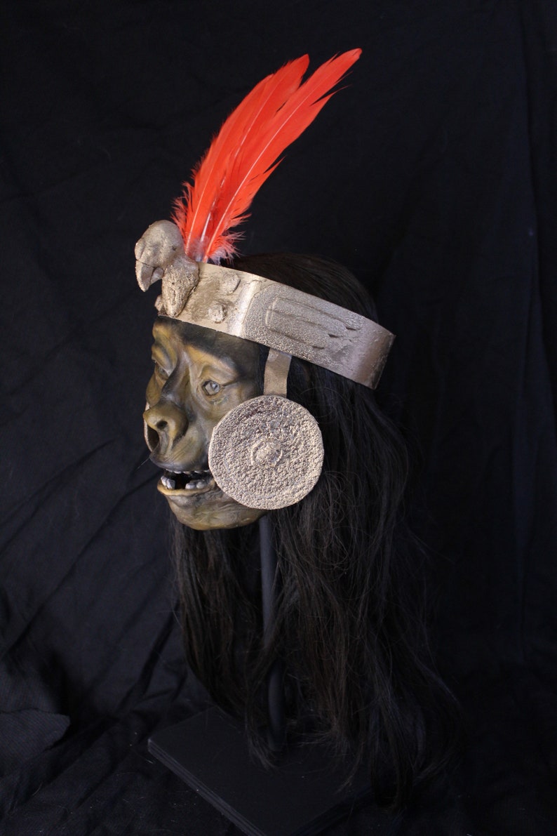 May include: A detailed, hand-crafted mask of a monkey wearing a silver headdress with red feathers. The mask has a realistic appearance with a textured, green face and long black hair.