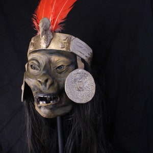 May include: A dark green and brown monkey mask with a gold and silver headdress and red feathers. The mask has a wide mouth with teeth and a long black wig.