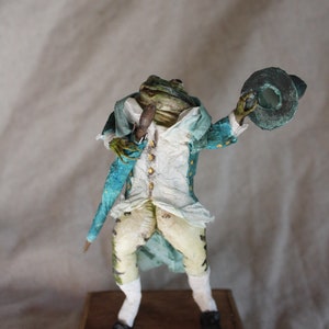 Może przedstawiać: Figurkę żaby w niebiesko-białej kurtce, białych spodniach i czarnym kapeluszu. Figurka trzyma laskę w prawej ręce i stoi na drewnianej podstawie.