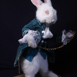 Könnte beinhalten: Ein weißes Kaninchen, das eine blaugrüne und weiße Weste mit einer goldenen Kette und einer Taschenuhr trägt. Das Kaninchen steht auf einem Holzsockel.