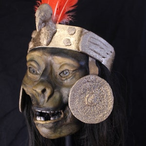 May include: A dark green and brown mask with a toothy grin, wearing a gold and silver headdress with a red feather. The mask has a round, gold disc on each side.