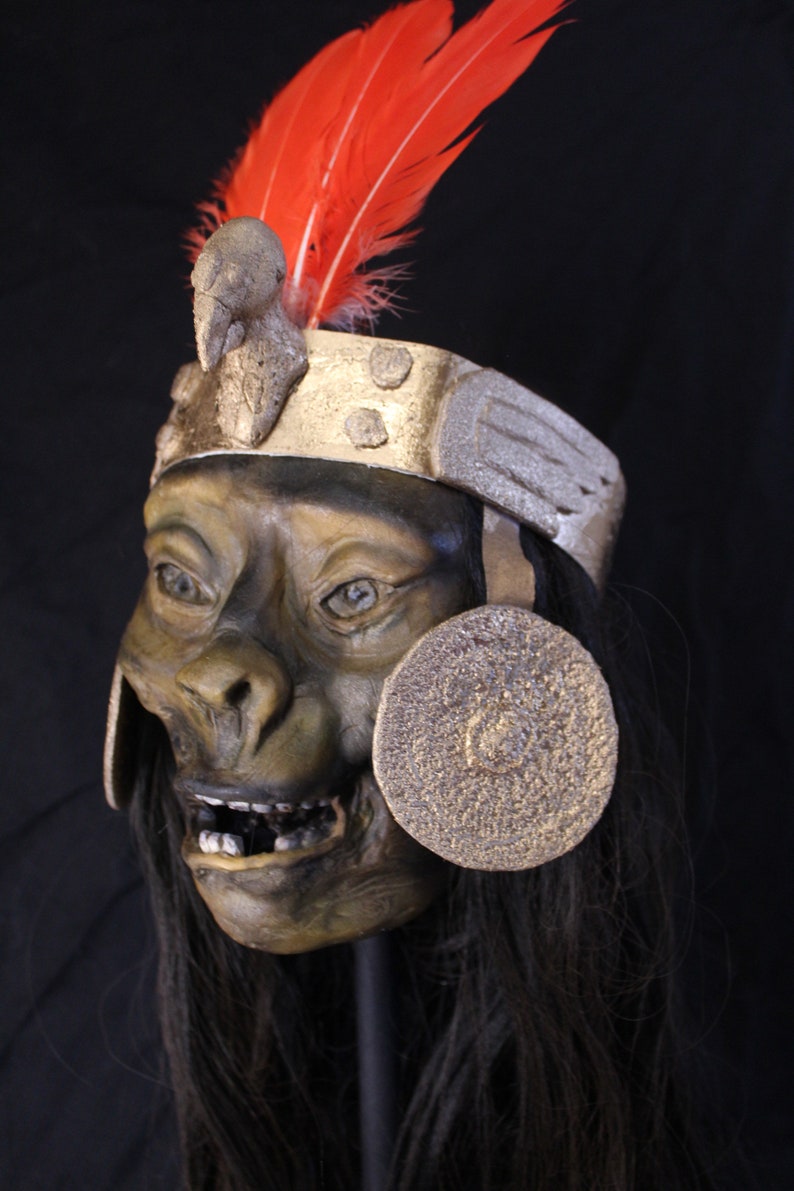 May include: A dark green and brown mask with a toothy grin, wearing a gold and silver headdress with a red feather. The mask has a round, gold disc on each side.