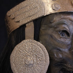 May include: A close-up of a dark-skinned, mummified head wearing a gold-colored headdress with a red feather. The headdress has a wing-like design and a round, gold-colored disc on the side of the head.