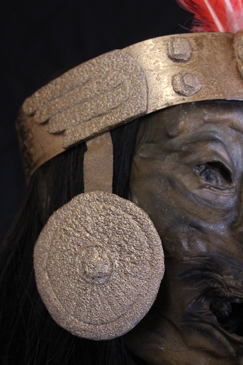 May include: A close-up of a dark-skinned, mummified head wearing a gold-colored headdress with a red feather. The headdress has a wing-like design and a round, gold-colored disc on the side of the head.